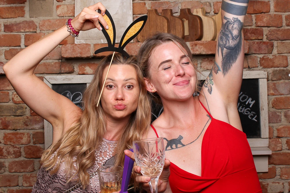 Zwei Frauen mit lustigen Fotobox-Accessoires und Gläsern in der Hand, die fröhlich in die Kamera lächeln.