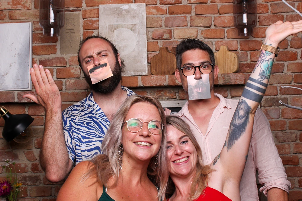 Vier Freunde posieren in einer Fotobox, zwei von ihnen halten witzige Gesichtsmasken hoch.